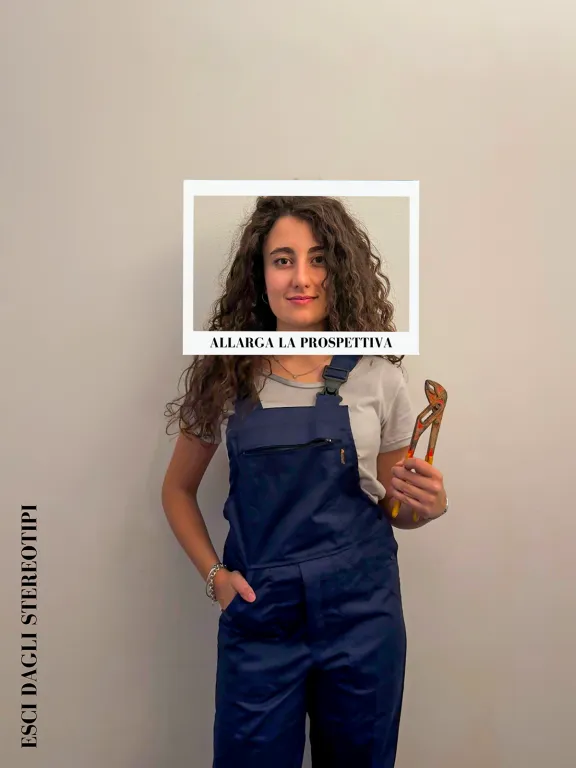 donna che al di fuori della cornice indossa una tuta da lavoro e tiene in mano una pinza