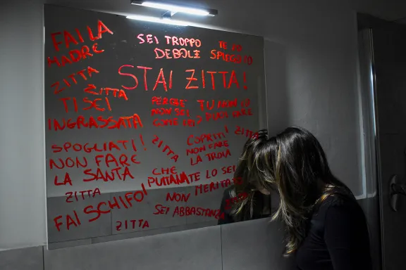 donna davanti a uno specchio che reca scritte come: "Stai zitta! Fai la madre. Sei troppo debole..."